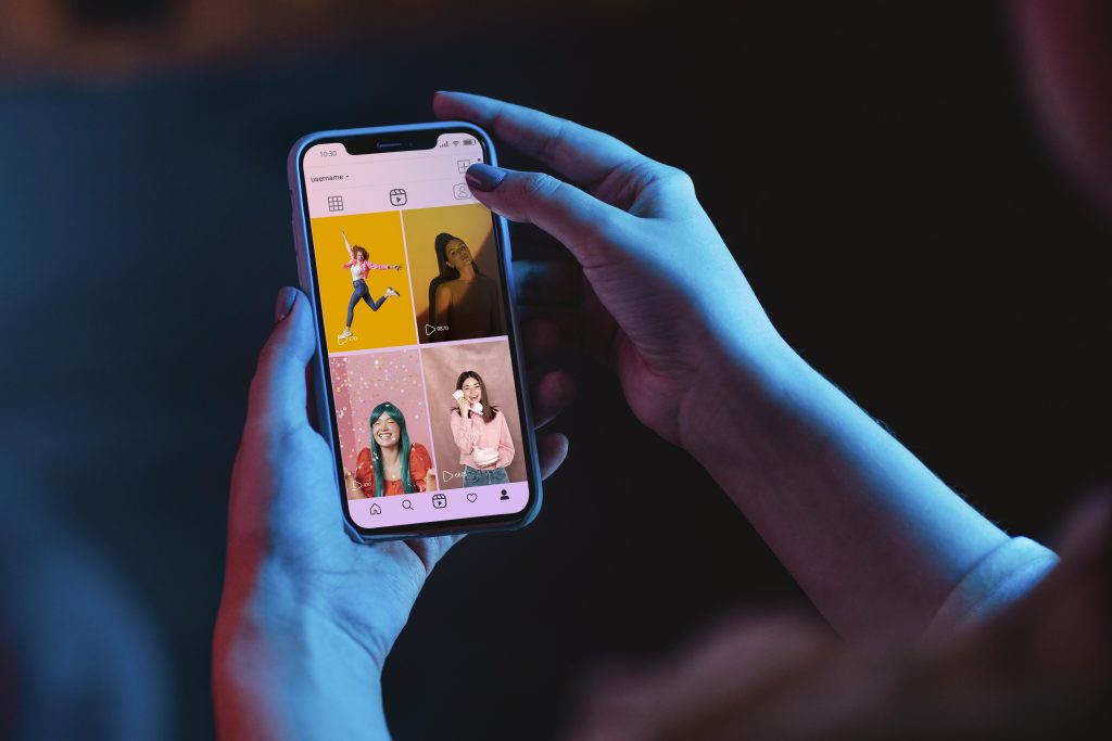 Mãos segurando um smartphone exibindo a interface do Instagram Reels, com vídeos de criadores de conteúdo em diferentes cenários e cores vibrantes. O fundo escuro e a iluminação azul destacam a tela do dispositivo.