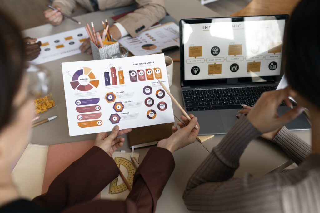 Pessoas em uma reunião de trabalho analisando gráficos e infográficos impressos e digitais. Duas pessoas em primeiro plano seguram um relatório impresso, enquanto uma tela de laptop exibe informações visuais. No fundo, outras pessoas participam da discussão em uma mesa com materiais de escritório.