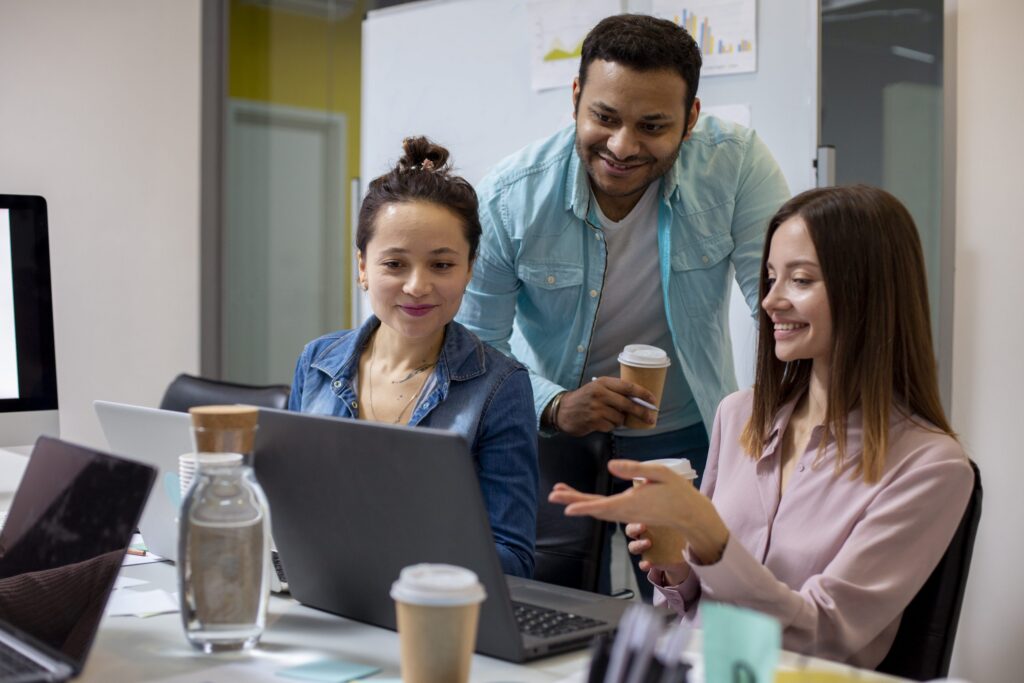 Três colegas diversos colaboram em torno de laptops em uma mesa de escritório, sorrindo e gesticulando enquanto discutem o conteúdo da tela, com foco no trabalho em equipe e na resolução de problemas.